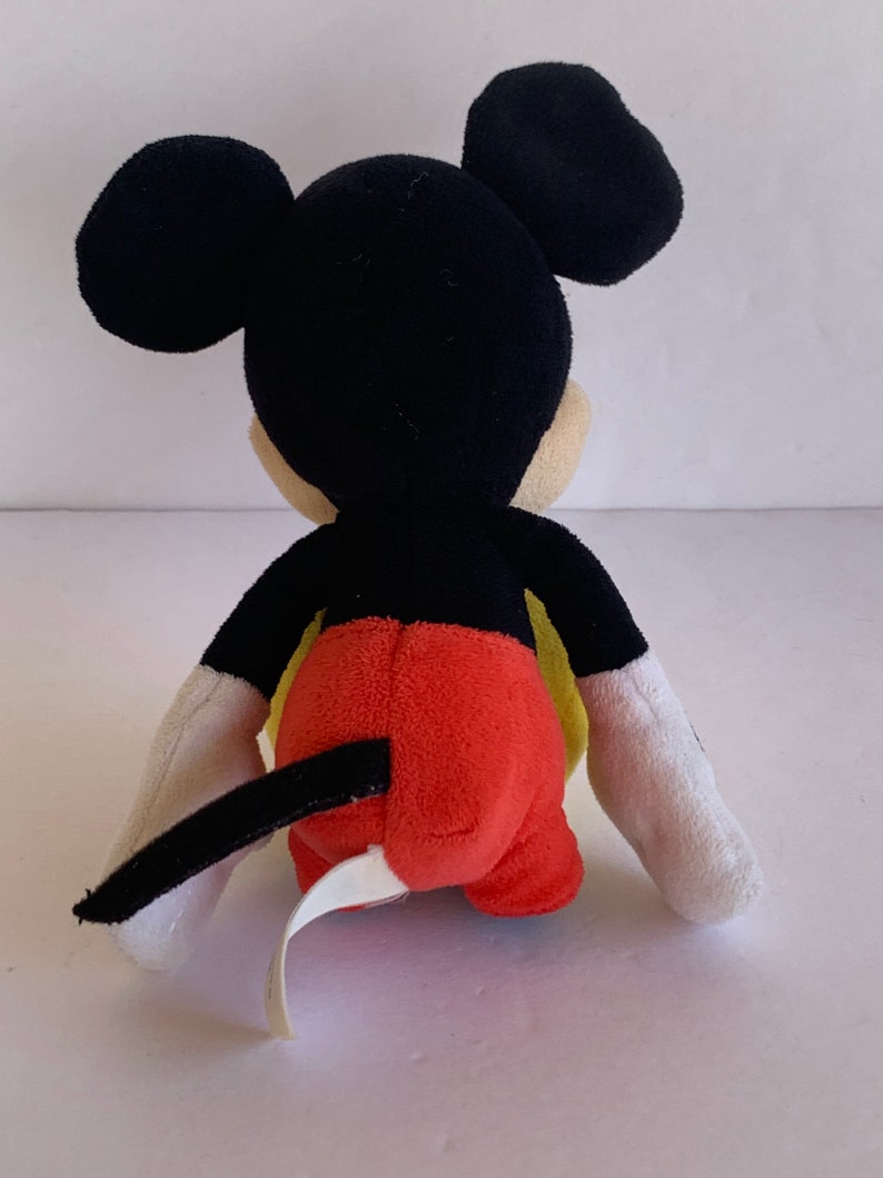Op de afbeelding: Een pluche Mickey Mouse-speelgoed, gezien van achteren, met zwarte oren, hoofd en staart. Het speelgoed heeft een rode onderkant, gele accenten en witte handschoenen. Een wit label is aan het speelgoed bevestigd.