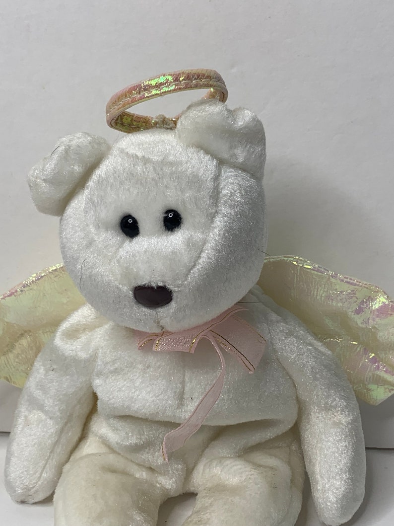 May include: White plush teddy bear with a pink ribbon and iridescent wings and halo. The bear is sitting with its wings spread out.