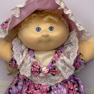 May include: A vintage doll with blonde hair and blue eyes, wearing a pink bonnet with lace trim. The doll is dressed in a floral print dress with a white lace collar and a pink bow.