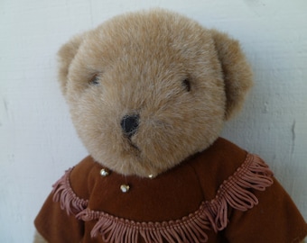 Peut inclure: Un ours en peluche avec une fourrure brun clair et une tenue marron foncé. La tenue a des franges et des accents dorés. Le nez de l'ours est noir et il a de petits yeux sombres.