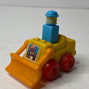 May include: A yellow toy bulldozer with a red front blade and red wheels. A blue toy person with a yellow face and a blue hat is sitting in the driver's seat. The bulldozer has a small window with a picture of a brown toy animal wearing a pink hat.
