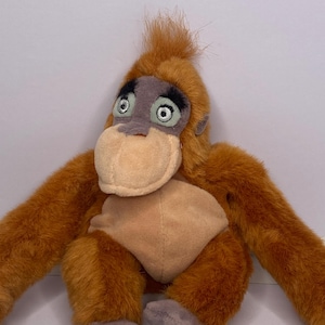 May include: A plush orangutan toy with orange fur, a tan face, and gray accents around the eyes and feet. The toy has long arms and a tuft of hair on its head. The toy is designed to resemble an ape.