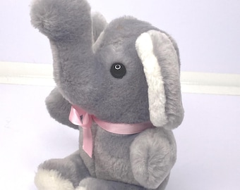 May include: A gray plush elephant toy with a pink ribbon around its neck. The elephant has white accents on its ears and feet, and a black eye. The toy is sitting on a white surface.