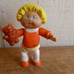 Op de afbeelding: Een plastic pop met blond haar, die een oranje shirt en een witte broek draagt. De pop houdt een bruine teddybeer vast.