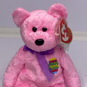 May include: A pink plush bear with a purple ribbon and an embroidered Easter egg design. The bear has black eyes and a red and white tag attached to its ear. A collectible toy.