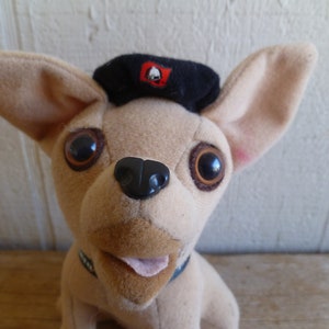 Puede incluir: Un chihuahua de peluche marrón que lleva una boina negra con una campana roja en la parte superior. El chihuahua tiene grandes ojos marrones y una nariz negra.