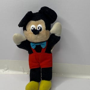 Könnte beinhalten: Ein schwarzes und braunes Plüsch-Mickey-Mouse-Spielzeug, das rote Shorts, eine blaue Fliege und gelbe Schuhe trägt. Das Spielzeug steht auf einer weißen Oberfläche.