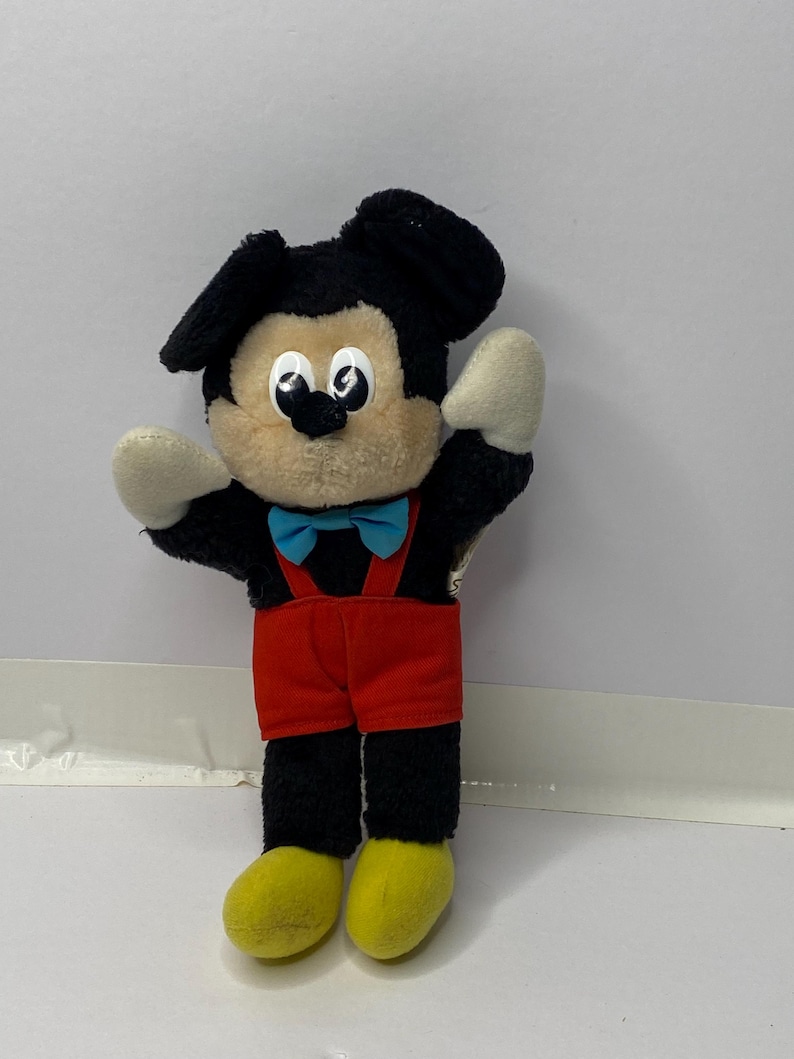 May include: A black and brown plush Mickey Mouse toy wearing red shorts, a blue bow tie, and yellow shoes. The toy is standing on a white surface.
