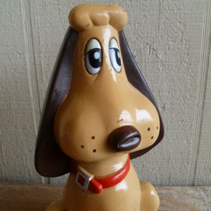 Może przedstawiać: Ceramiczna figurka psa w kolorze brązowym i beżowym z dużymi brązowymi uszami i czerwonym kołnierzem. Pies ma głupi wyraz twarzy z dużymi oczami i małym czarnym nosem.