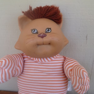 May include: A vintage cat doll with a peach-coloured face and brown hair. The doll wears a striped orange and white shirt. The cat doll has a smiling expression and is a collectible item.