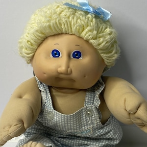 Może przedstawiać: Lalka Cabbage Patch Kid z blondą wełnianą czupryną, ubrana w żółty, dziergany na szydełku kapelusz z niebieską kokardką i niebiesko-białe spodnie ogrodniczki.