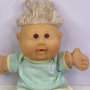 May include: A doll with light skin, brown eyes, and messy blonde hair. The doll wears a light green short-sleeved shirt with a white heart-shaped logo containing the letters CPK, paired with cream-colored shorts. The doll has a soft fabric body.