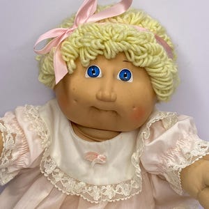 May include: A vintage doll with a soft, vinyl face, blue eyes, and yarn hair. The doll wears a pink dress with lace trim and a matching pink bow in its hair. The doll has rosy cheeks and a sweet expression.