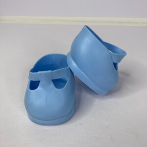 May include: A pair of pale blue Mary Jane shoes. The shoes feature a rounded toe and a strap that fastens over the foot. They are made from a soft, flexible material and are designed for children's wear.