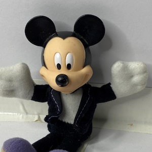 May include: A plush Mickey Mouse toy wearing a black tuxedo with a white shirt and purple shoes. The toy has a black nose and large black ears.
