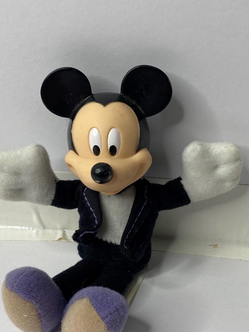 May include: A plush Mickey Mouse toy wearing a black tuxedo with a white shirt and purple shoes. The toy has a black nose and large black ears.