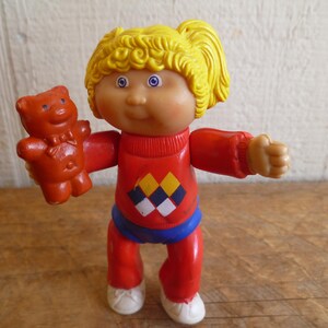 Op de afbeelding: Een plastic pop met blond haar die een rode trui draagt met een zwart en geel ruitpatroon, een blauwe broek en witte schoenen. De pop houdt een bruine teddybeer vast.