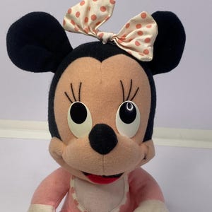 Peut inclure: Poup&eacute;e Minnie Mouse en peluche avec des oreilles noires, un visage rose et une tenue rose assortie. La poup&eacute;e a un n&oelig;ud cr&egrave;me &agrave; pois p&ecirc;che et de grands yeux ronds. Un bavoir blanc &agrave; bord festonn&eacute; compl&egrave;te le look.