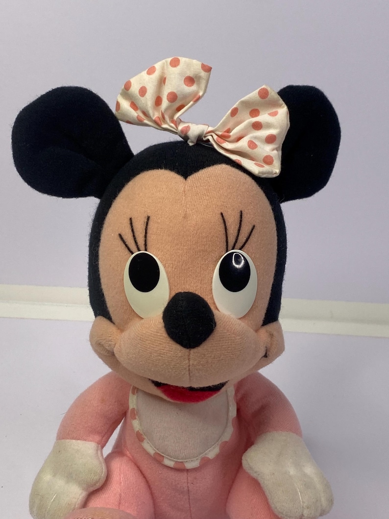 Peut inclure: Poup&eacute;e Minnie Mouse en peluche avec des oreilles noires, un visage rose et une tenue rose assortie. La poup&eacute;e a un n&oelig;ud cr&egrave;me &agrave; pois p&ecirc;che et de grands yeux ronds. Un bavoir blanc &agrave; bord festonn&eacute; compl&egrave;te le look.