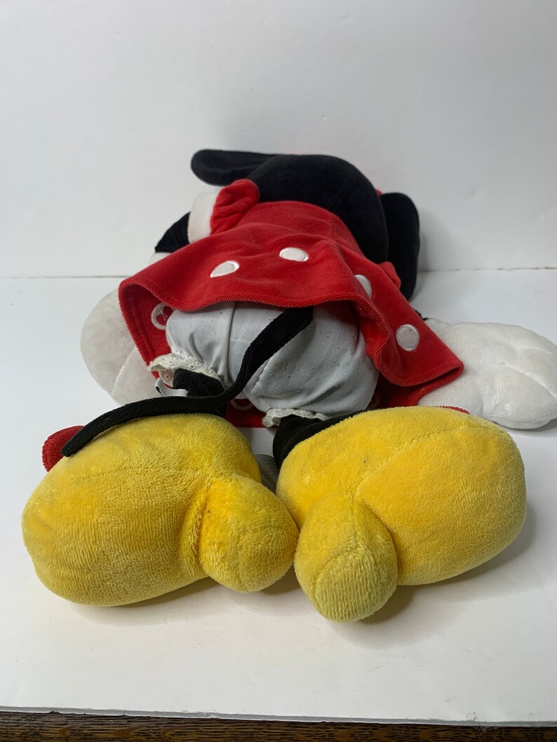 Pu&ograve; includere: Un giocattolo di peluche rosso e nero di Minnie Mouse. Il giocattolo &egrave; sdraiato sulla schiena con le braccia e le gambe distese. Il giocattolo indossa un vestito rosso con pois bianchi e un fiocco nero.