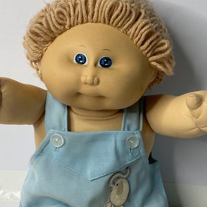 May include: A Cabbage Patch Kid doll with blonde yarn hair, blue eyes, and a light blue overall outfit with an embroidered elephant design.