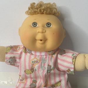 Könnte beinhalten: Eine Cabbage Patch Kid Puppe mit blonden Locken, die einen rosa-weiß gestreiften Schlafanzug mit Teddybär-Motiven trägt. Der Schlafanzug hat den Text "CPK" auf der Vorderseite.