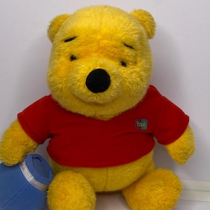 Könnte beinhalten: Ein gelber Plüschbär mit rotem T-Shirt und dem Wort "Pooh". Der Bär hat eine schwarze Nase und hält einen blauen Honigtopf mit silbernem Band.