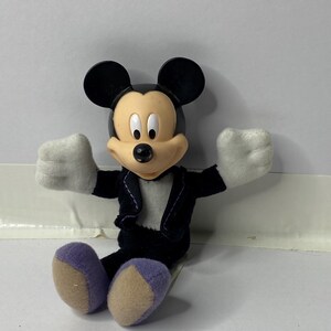 May include: A plush Mickey Mouse toy wearing a black tuxedo with a white shirt and purple shoes. The toy is sitting with its arms outstretched.