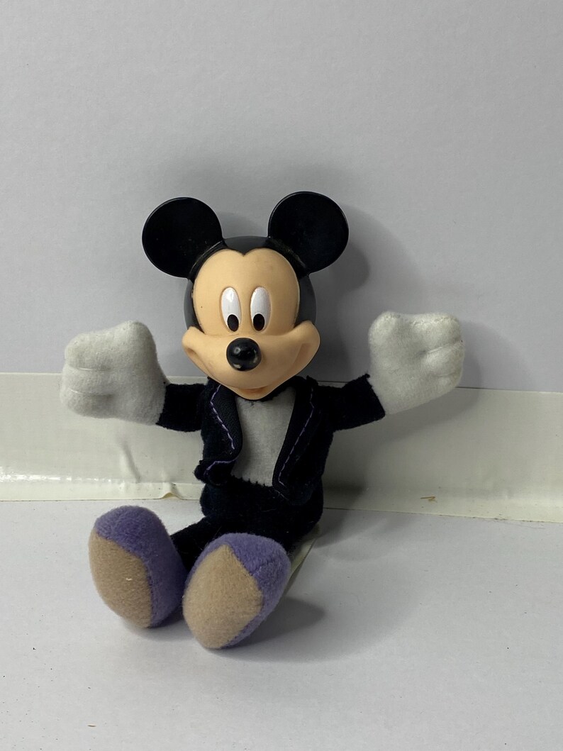 May include: A plush Mickey Mouse toy wearing a black tuxedo with a white shirt and purple shoes. The toy is sitting with its arms outstretched.