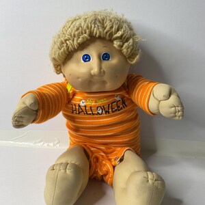 Könnte beinhalten: Eine Cabbage Patch Kid Puppe, die einen orange-weiß gestreiften Strampler mit dem Text "My first HALLOWEEN" auf der Vorderseite trägt. Die Puppe hat blondes Garnhaar und blaue Augen.