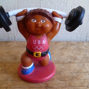 Könnte beinhalten: Eine braune Cabbage Patch Kid-Puppe, die ein rot-weiß-blaues USA-Olympia-Outfit trägt und eine schwarze Langhantel hochhebt.