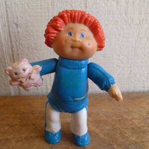 Op de afbeelding: Een vintage Cabbage Patch Kids pop met rood krullend haar, blauwe ogen en een roze teint. De pop draagt een blauwe outfit met een witte broek en blauwe schoenen, en houdt een kleine roze teddybeer vast. Een klassiek verzamelobject.