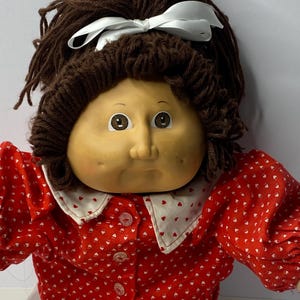 Puede incluir: Una muñeca Cabbage Patch Kid con pelo de lana marrón, un lazo blanco y una camisa con estampado de corazones rojos y blancos.