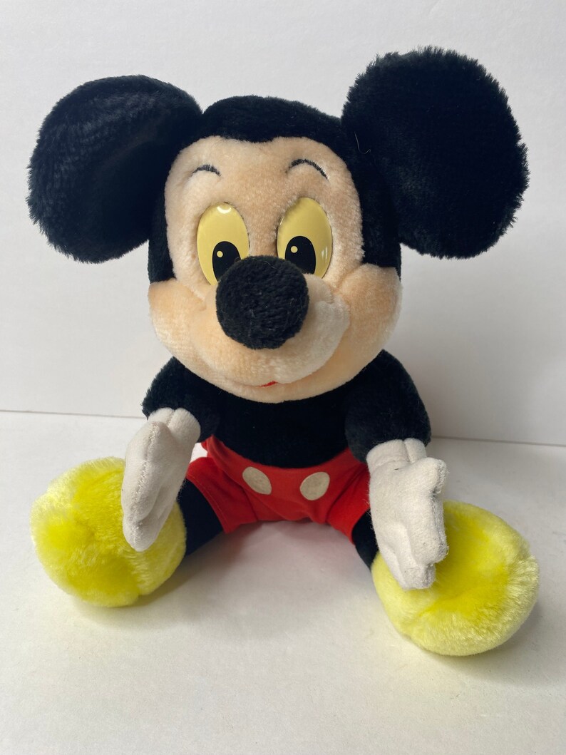 May include: A plush Mickey Mouse toy sitting with his legs crossed. He has black fur, large ears, a red and white polka dot shorts, and yellow shoes.