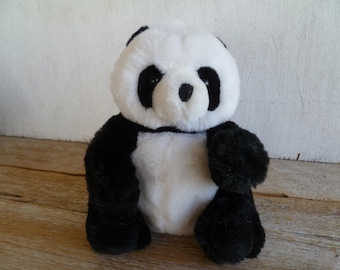 May include: A plush panda bear with black and white fur. The panda has black fur around its eyes, ears, and limbs, with a white body and face. The stuffed animal is sitting on a wooden surface.