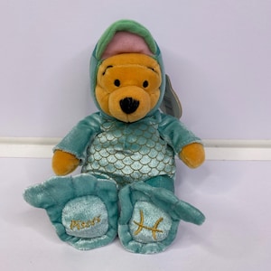 Könnte beinhalten: Ein Winnie Puuh Plüschtier in einem türkisfarbenen Fischkostüm mit Schuppenmuster am Oberkörper und Kapuze. Die Füße sind mit goldener Stickerei versehen, wobei auf dem linken Fuß das Wort Pisces und auf dem rechten das Sternzeichen Fische steht.