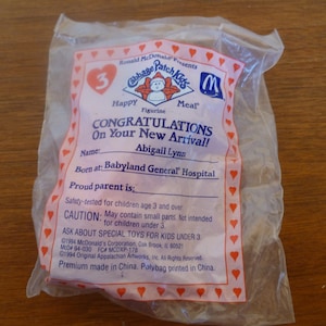Puede incluir: Una bolsa de plástico con una etiqueta rosa y blanca que dice "Ronald McDonald Presenta Cabbage Patch Kids Happy Meal". La etiqueta también tiene una imagen de una muñeca Cabbage Patch Kid y el número 3. La etiqueta dice "¡Felicidades por tu nueva llegada!" y tiene un espacio para escribir el nombre del bebé, el lugar de nacimiento y el nombre del orgulloso padre. La etiqueta también tiene una advertencia que dice "Probado para la seguridad de los niños de 3 años en adelante. PRECAUCIÓN: Puede contener piezas pequeñas. No apto para niños menores de 3 años. Pregunte por los juguetes especiales para niños menores de 3 años."