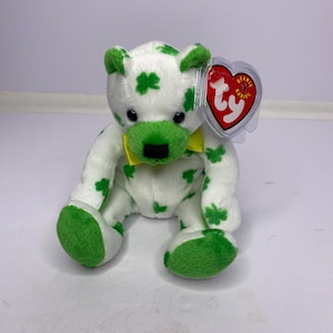 May include: A white plush bear with green shamrock patterns, green accents on the ears, paws, and muzzle, and a yellow bow. The bear has a black nose and eyes, and a red heart-shaped tag that reads "Beanie Babies".