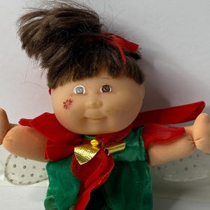 May include: A Cabbage Patch Kid doll dressed as a Christmas elf. The doll has brown hair, a green dress, red cape, and gold trim. The doll has one blue eye and one brown eye.
