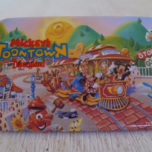 May include: A rectangular magnet featuring a colorful illustration of Mickey's Toontown at Disneyland. The image includes a train with Mickey Mouse, Minnie Mouse, Donald Duck, and other characters. The background shows a sunny sky, green hills, and buildings.