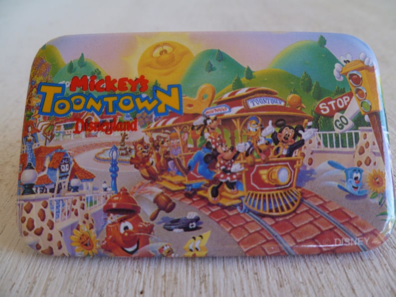 May include: A rectangular magnet featuring a colorful illustration of Mickey's Toontown at Disneyland. The image includes a train with Mickey Mouse, Minnie Mouse, Donald Duck, and other characters. The background shows a sunny sky, green hills, and buildings.