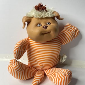 May include: A plush toy dog wearing orange and white striped clothing. The dog has a brown and white furry head with large eyes and a brown nose.