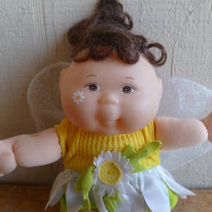 May include: A doll with brown hair wearing a yellow dress with a white daisy on the front. The doll has a white and silver fairy wings.