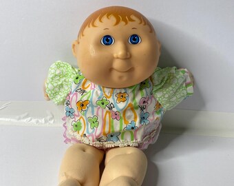 Puede incluir: Una muñeca Cabbage Patch Kid con un vestido de estampado floral verde y blanco, ojos azules y una peluca marrón claro.