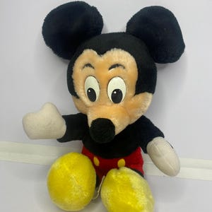 May include: A vintage Mickey Mouse soft toy. The plush has black ears, a black head, and a tan face. It features white hands, red shorts, yellow feet, and large eyes. The toy is in a sitting position.