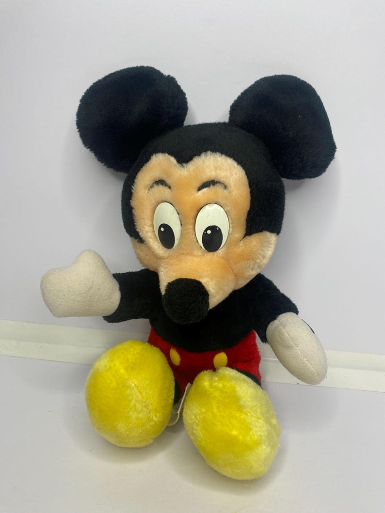 May include: A vintage Mickey Mouse soft toy. The plush has black ears, a black head, and a tan face. It features white hands, red shorts, yellow feet, and large eyes. The toy is in a sitting position.