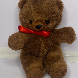 Puede incluir: Un oso de peluche marrón con una cinta roja alrededor del cuello. El juguete de peluche tiene una textura suave y peluda y ojos negros. Un juguete clásico para niños.