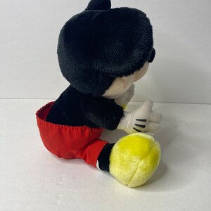 May include: A plush Mickey Mouse toy sitting with red shorts and yellow shoes.