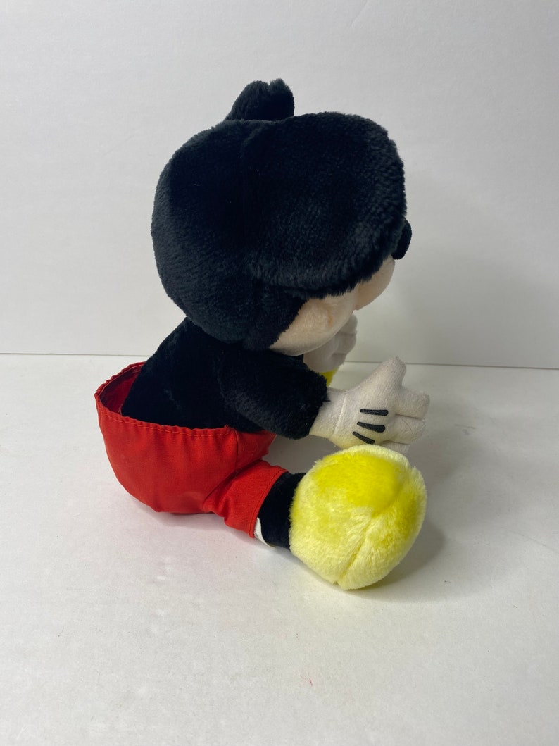 May include: A plush Mickey Mouse toy sitting with red shorts and yellow shoes.