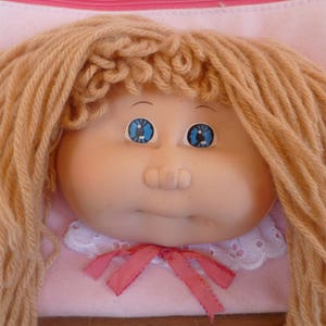 May include: A doll with long, blonde yarn hair, blue eyes, and a pink dress with a white lace collar and a pink bow.